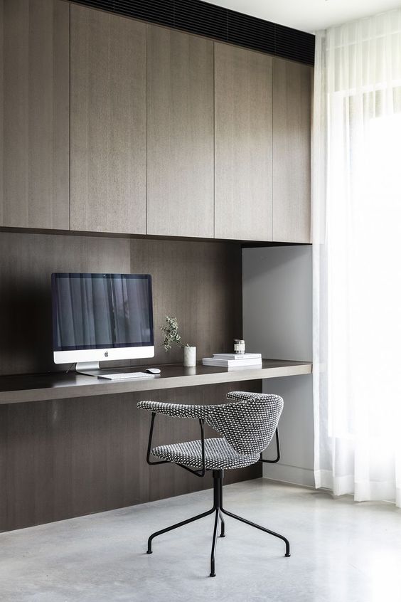 A refined minimalist home office with dark stained sleek cabinets, a built in desk, a grey chair and a large window for more natural light