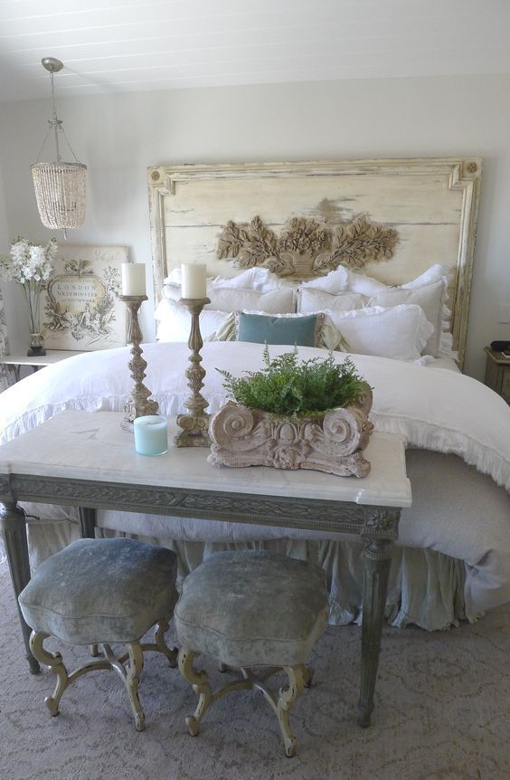 a shabby chic bedroom with a neutral wooden bed, a table and grey stools, a bed chandelier and candles