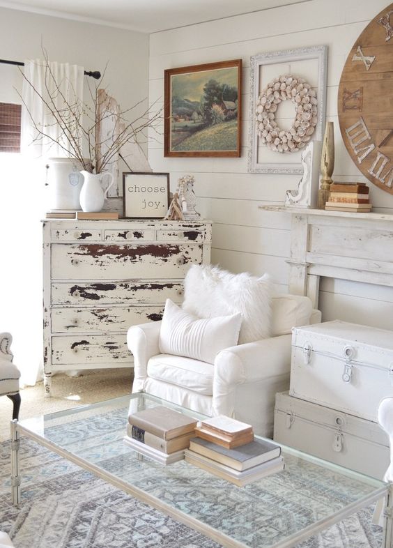 a shabby chic living room with a faux fireplace, white chests, white chairs, a shabby sideboard and a glass table