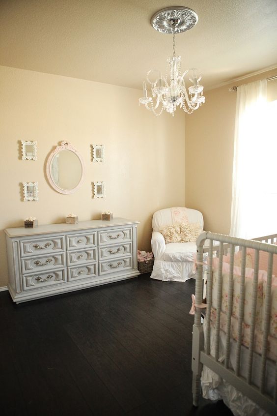 a shabby chic nursery with tan walls, elegant grey furniture, a white chair, a pretty vintage chandelier and a gallery wall with mirrors
