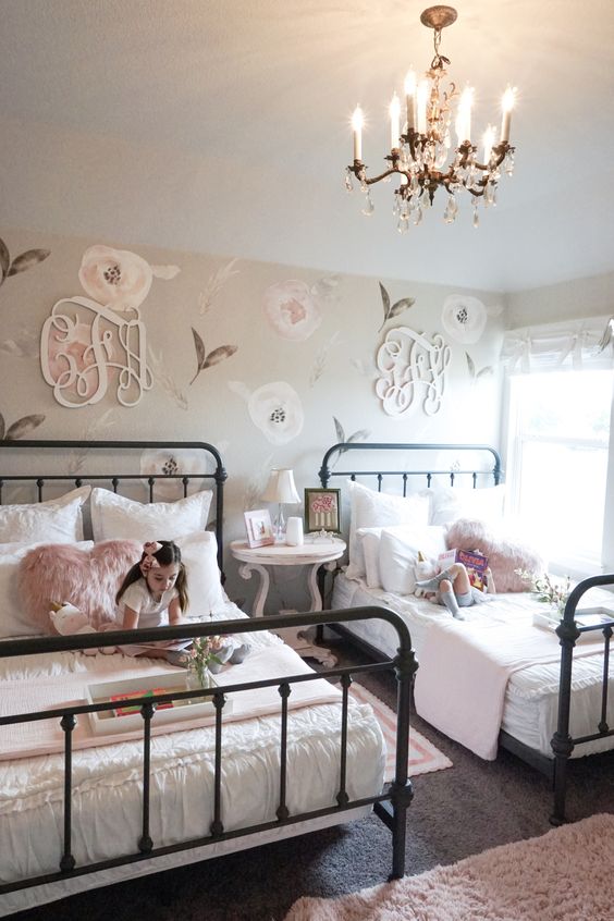 a shabby chic shared bedroom with a floral statement wall, metal beds, pink bedding, a refined nightstand and a crystal chandelier