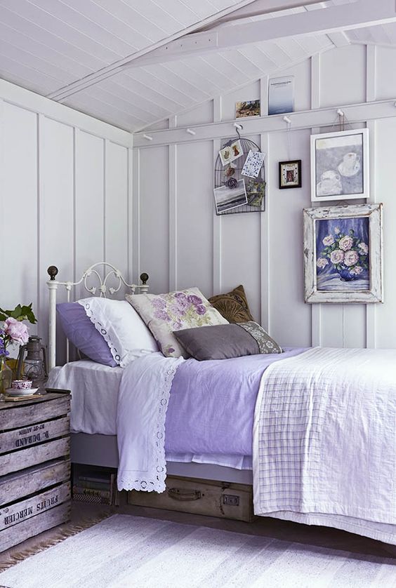 a shabby farmhouse bedroom in white, with a forged bed, a crate nightstand, some vintage suitcases and a gallery wall