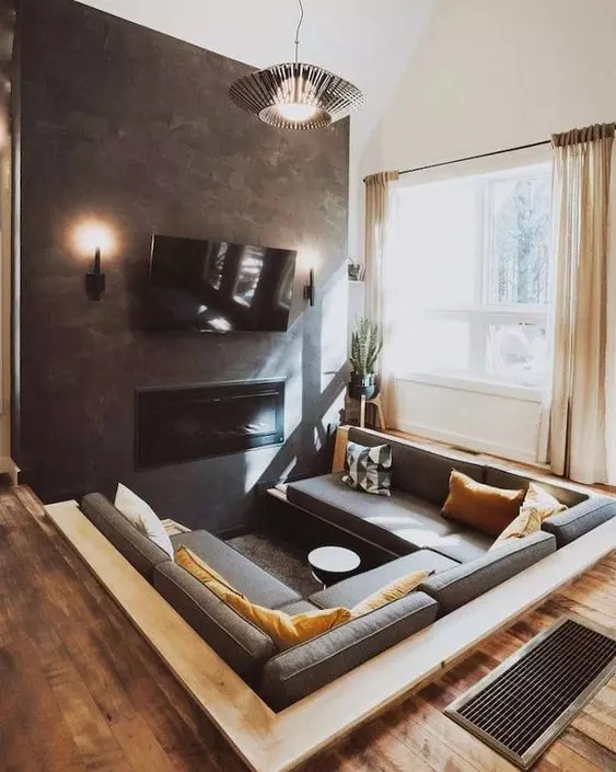 A small yet cozy modern conversation pit with a grey sofa that takes three sides, bold pillows, a built in fireplace and a TV