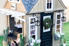 a stylish farmhouse playhouse with a black door, white window frames, a built-in bench, potted blooms and cacti