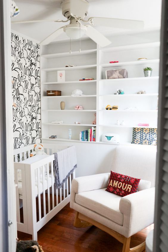 A tiny neutral nursery with a bunny accent wall, built in shelves, a white crib and a rocker plus some bright textiles