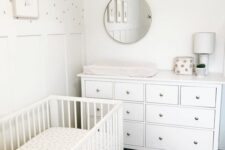a tiny neutral nursery with a white crib and a dresser, layered rugs, a polka dot accent wall and a round mirror