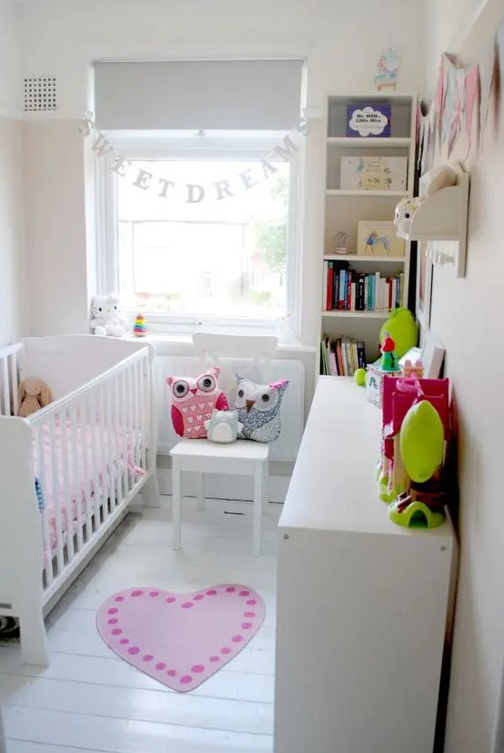 A tiny neutral nursery with white furniture, a built in bookshelf and a dresser, a banner, some bright textiles and colorful toys