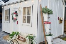 a white farmhouse playhouse with lights, greenery and blooms, a ladder with plants in buckets and a wreath is super cute