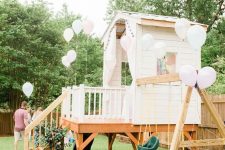 a white kids’ playhouse with a porch, a ladder, swings and white balloons is a lovely idea for your garden