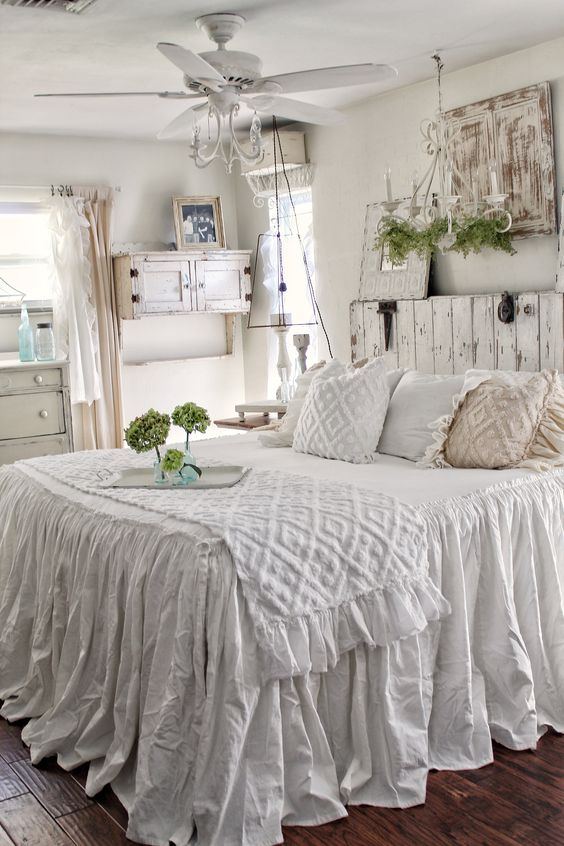 a white shabby chic bedroom with a wooden bed and a wooden screen, a cabinet, a white fan and a moss chandelier and greenery