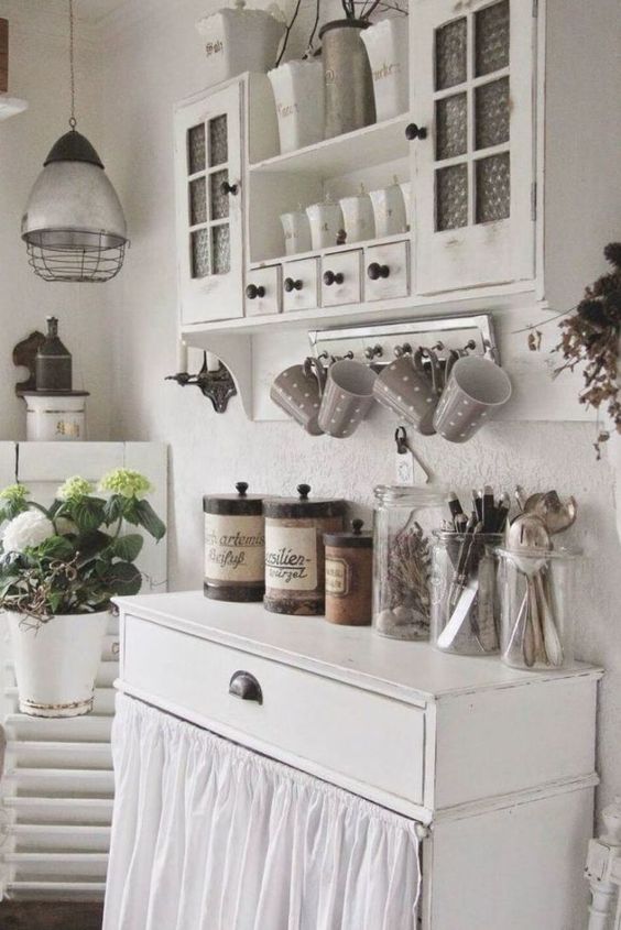 a white shabby chic kitchen with vintage furniture, a cabinet with a curtain, potted blooms, a metal pendant lamp