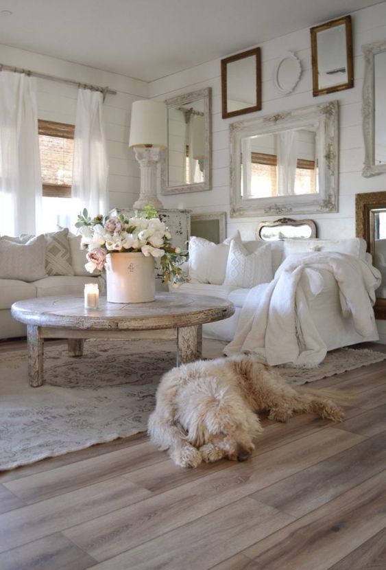 a white shabby chic living room with a gallery wall of mirrors, white furniture, a shabby table and lamps and blooms