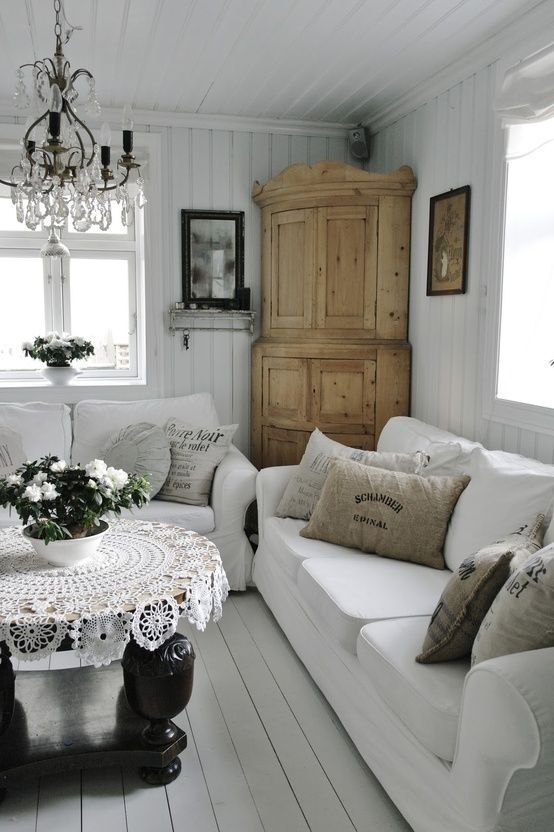 a white vintage to shabby chic living room with white sofas, a wooden wardrobe, a crystal chandelier, a black table, a crochet tablecloth and blooms