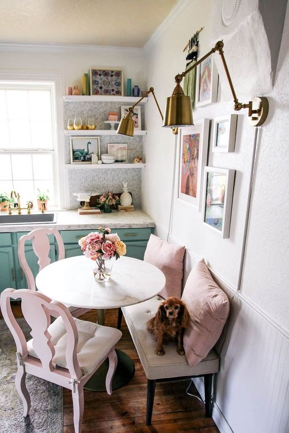 a cute small nook with a neutral bench, pink chairs and pillows, a round table and a lovely bright gallery wlal