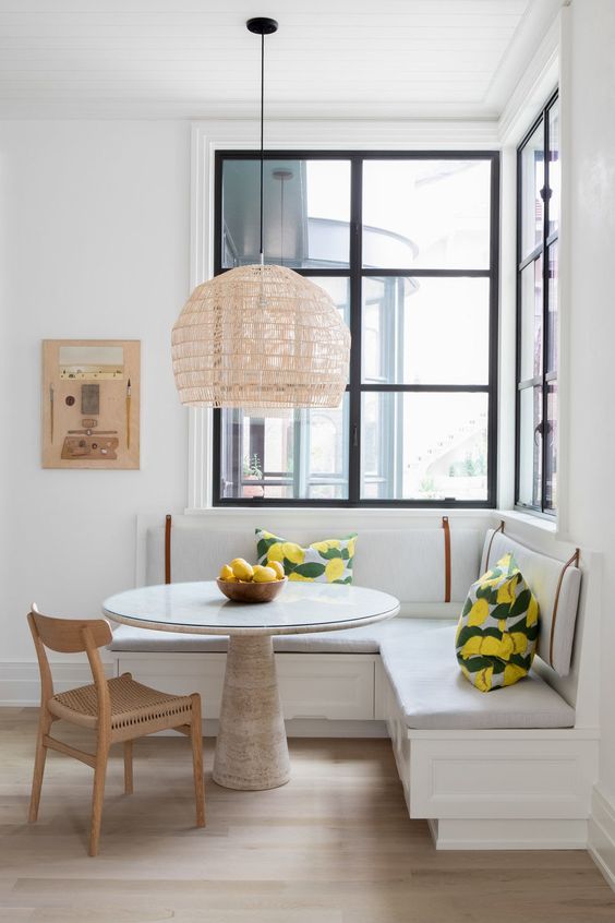 A light filled dining area by the windows, with a built in corner bench, a round table, a woven chair and a pendant lamp