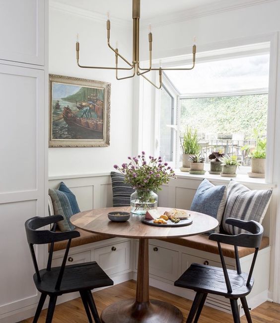 a lovely farmhouse dining nook with a built-in corner bench, printed pillows, a round table, black chairs and a gidled chandelier