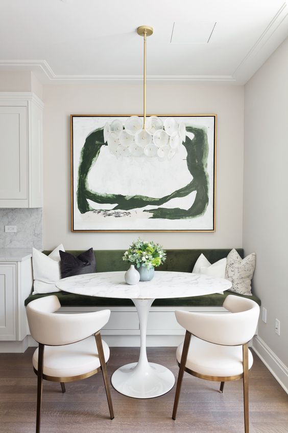 a small and elegant dining nook with a green bench, a round table, creamy rounded chairs, a bold artwork and a cool chandelier