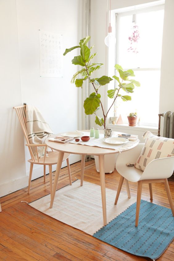 an airy small dining zone with a round table, stained and white chairs, potted plant, a hanging bulb and some printed textiles