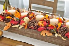 a bright fall centerpiece of a wooden box, fall leaves, acorns, gourds and pillar candles in glasses