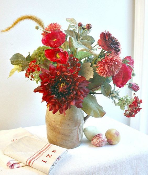 a bright fall centerpiece of red and burgundy blooms, greenery in a vintage jug is a bold decoration
