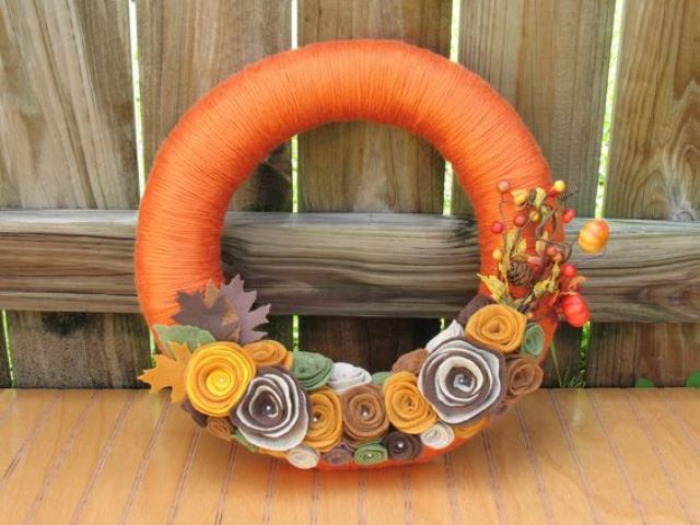 a bright orange yarn wreath decorated with fabric flowers, leaves, berries and dried blooms