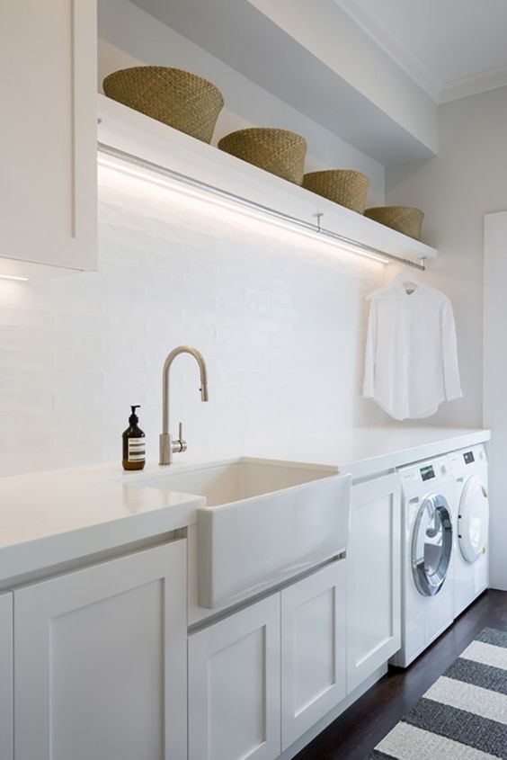 A clean neutral laundry room with white shaker style cabinets, white countertops, a square sink, baskets for storage and built in lights