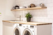 a clean white laundry with square tiles, an open shelf, a washing machine and a dryer, a butcherblock countertop and a potted plant
