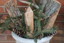 a concrete planter with pinecones, fir branches, firewood and lights is a cool outdoor decoration