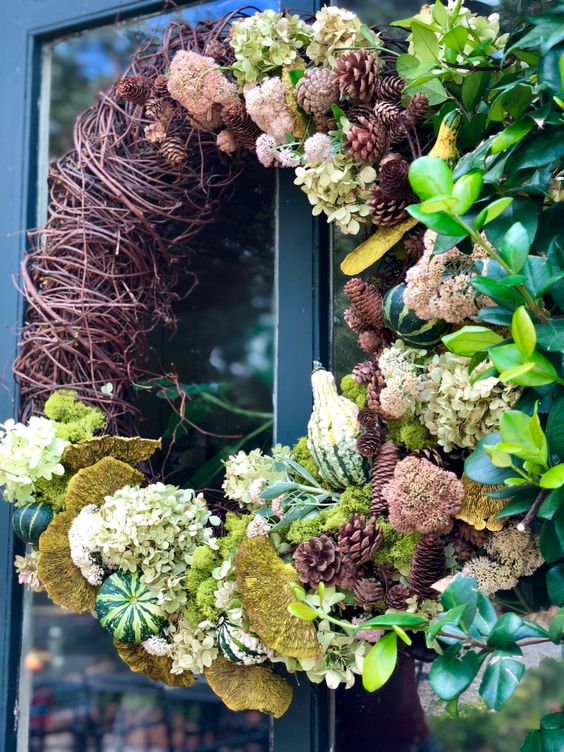 A creative fall wreath of white hydrangeas, vine, pinecones, greenery, gourds, mushrooms and greenery looks very eye catchy