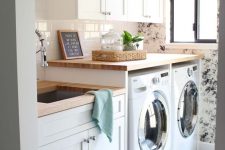a lovely laundry with a floral accent wall, white shaker style cabinets, butcherblock countertops and a sink, a basket for storage