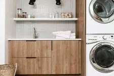 a modern laundry with grey skinny tiles, sleek plywood cabinets, a washing machine and a dryer, a jute rug and black sconces