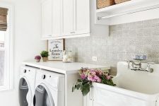 a modern neutral laundry with white shaker cabinets, a large sink, a vintage faucet, baskets for storage and a washing machine and a dryer