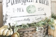 a shabby chic sign with black letters and a green pumpkin placed on a basket with greenery and surrounded by pumpkins