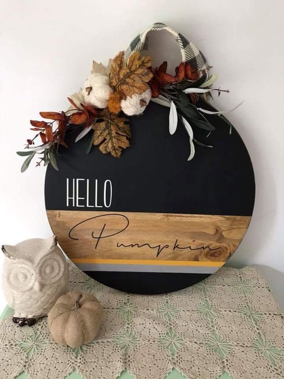 a stylish round fall sign done with staining and black paint, topped with leaves, yarn pumpkins and plaid ribbon
