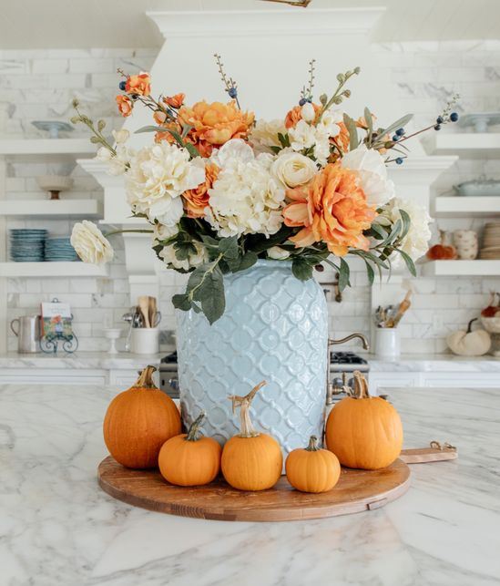 an elegant white and orange faux flower arrangement in a blue vase and orange pumpkins around it (designed by Holly Jolley)