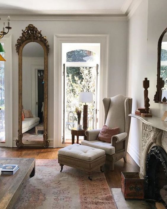 a French country living room with a tall mirror in a carved wooden frame, a neutral antique chair with a footrest, a vintage fireplace