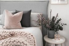 a Scandinavian bedroom with a grey upholstered bed, pink and grey pillows and a pink knit blanket, a grey planter with greenery