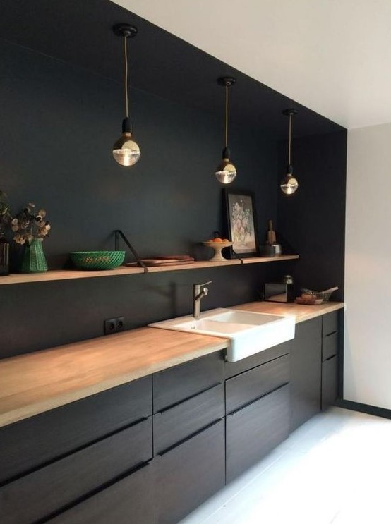 A black kitchen with light colored wood countertops, pendant lamps and open shelving over the cabinets
