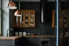 a black kitchen with matte cabinets, rich-stained upper ones, a black hood, copper pendant lamps and a black and white tile floor