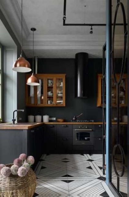 A black kitchen with matte cabinets, rich stained upper ones, a black hood, copper pendant lamps and a black and white tile floor