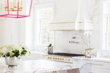 a bold glam kitchen with white cabinetry, gold handles and edging, a hot pink chandelier and acrylic stools with pink cushions