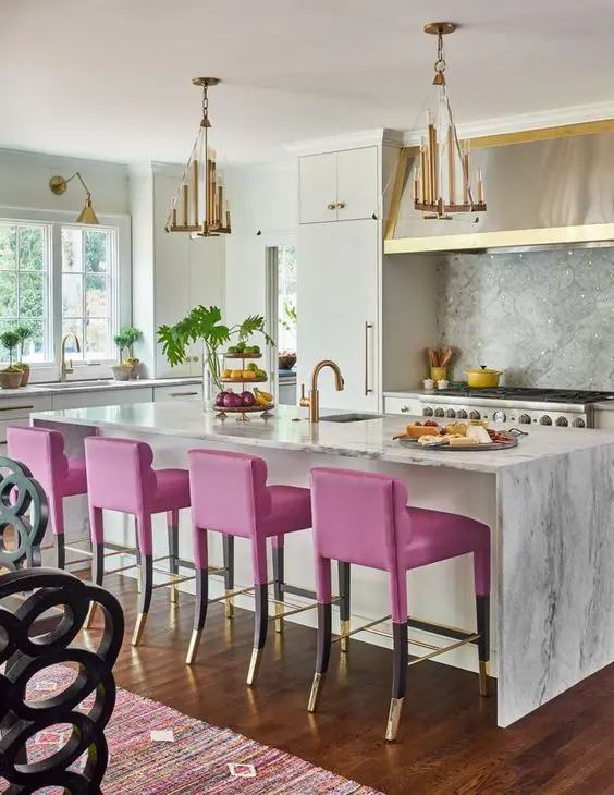 a bold glam kitchen with white cabinets, a grye backsplash and countertops, a shiny hood and metallic chandeliers, hot pink stools and gold fixtures