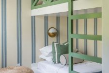 a bright kids’ room with striped walls, a green bunk bed, neutral and navy bedding, a jute pouf, a basket and toys