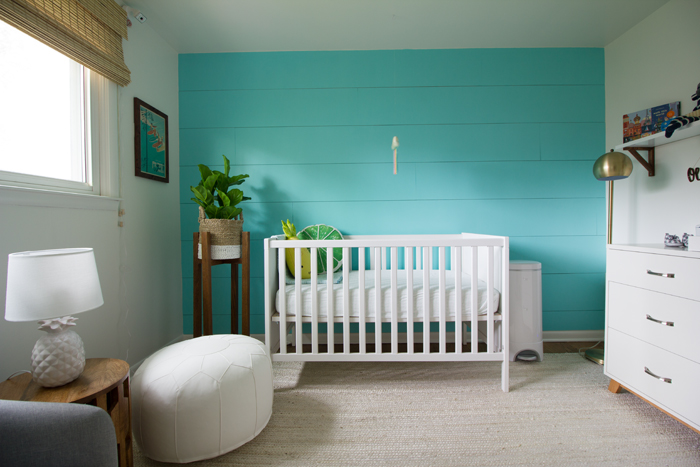 a chic and bright nursery with a statement turquoise wall, a white IKEA Sundvik crib and leather ottoman, wicker shades and a potted plant