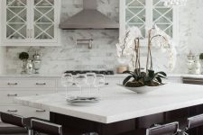 a chic glam kitchen with white cabinets and a chocolate brown kitchen island, crystal chandeliers, elegant curved stools