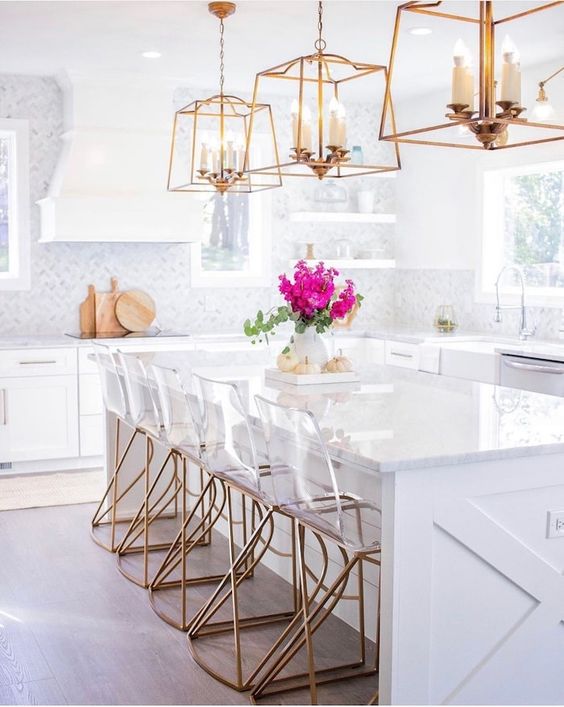 a contemporary glam kitchen with white cabinetry, white marble tiles, glam brass chandeliers and acryl and brass chairs