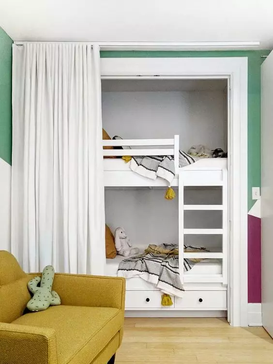 A cozy and bright kids' room with white built in bunk beds, neutral bedding, a mustard chair and a curtain to get some privacy