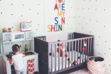 a cute pastel nursery with a dark stained Sundvik crib, colorful touches and an artwork, a play kitchen and polka dot walls