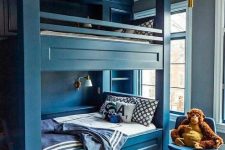 a deep blue kids’ room with built-in bunk beds and navy and white bedding, with printed pillows and blankets, a chair and a footrest
