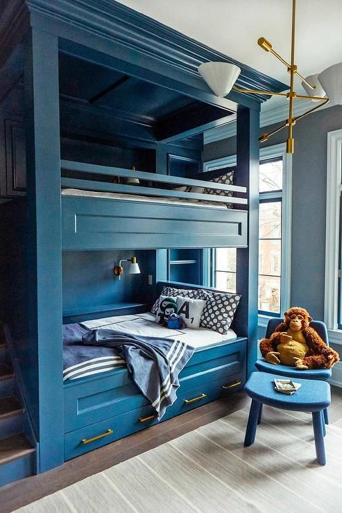 A deep blue kids' room with built in bunk beds and navy and white bedding, with printed pillows and blankets, a chair and a footrest
